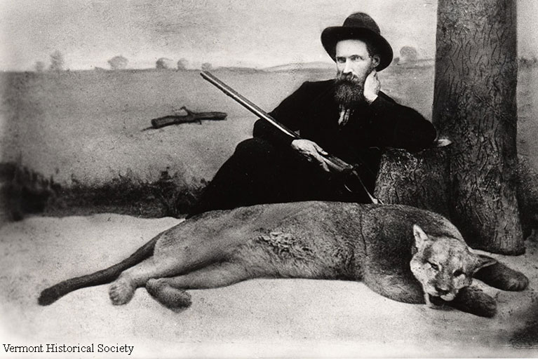 Man from the 1880s cradling a gun leaning against a fake tree with a dead panther in front of him